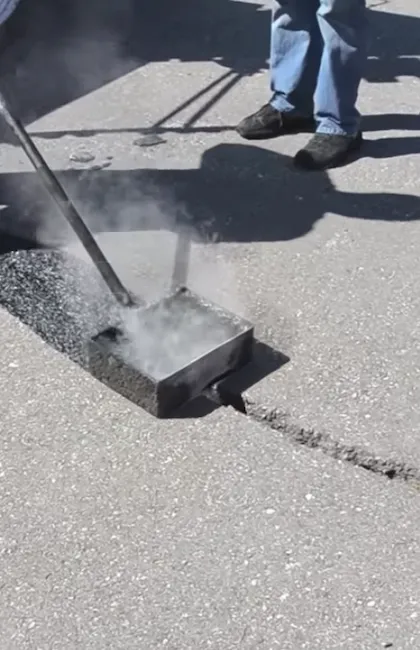 Une vue en plongée montre un ouvrier utilisant un applicateur de scellant chauffé pour réparer une fissure dans le pavage d'asphalte. De la vapeur ou de la fumée s'échappe du boîtier métallique rectangulaire alors qu'il est traîné le long de la fissure, laissant derrière lui une bande de bitume noir frais. On aperçoit les jambes de l'ouvrier en jeans bleus et chaussures de sécurité foncées sur le côté.