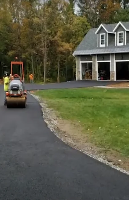 Un ouvrier en gilet jaune utilise un petit rouleau compresseur sur une nouvelle allée en asphalte devant une maison.